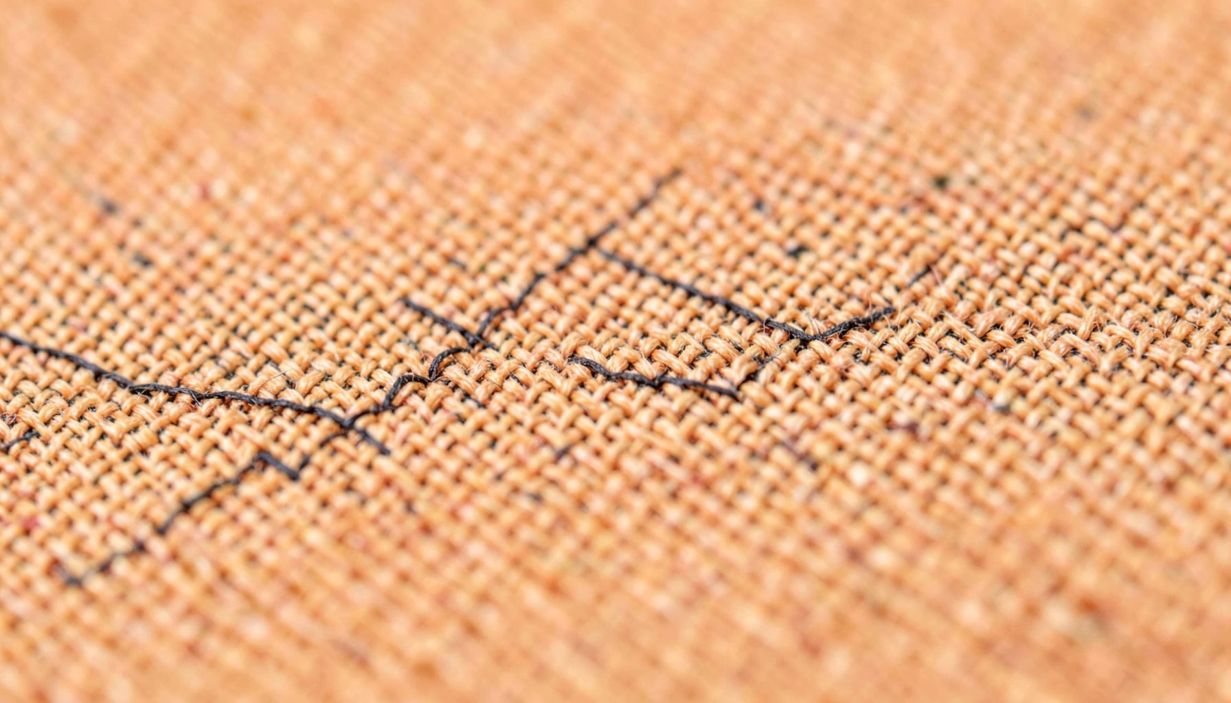 Close-up of woven tan fabric texture with fine charcoal stitching, calm and tactile in mood