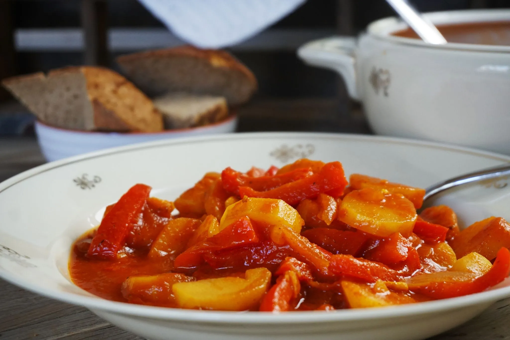 Close-up of a bowl of stewed vegetables in red sauce with bread and soup in background, warm red and orange tones, cozy