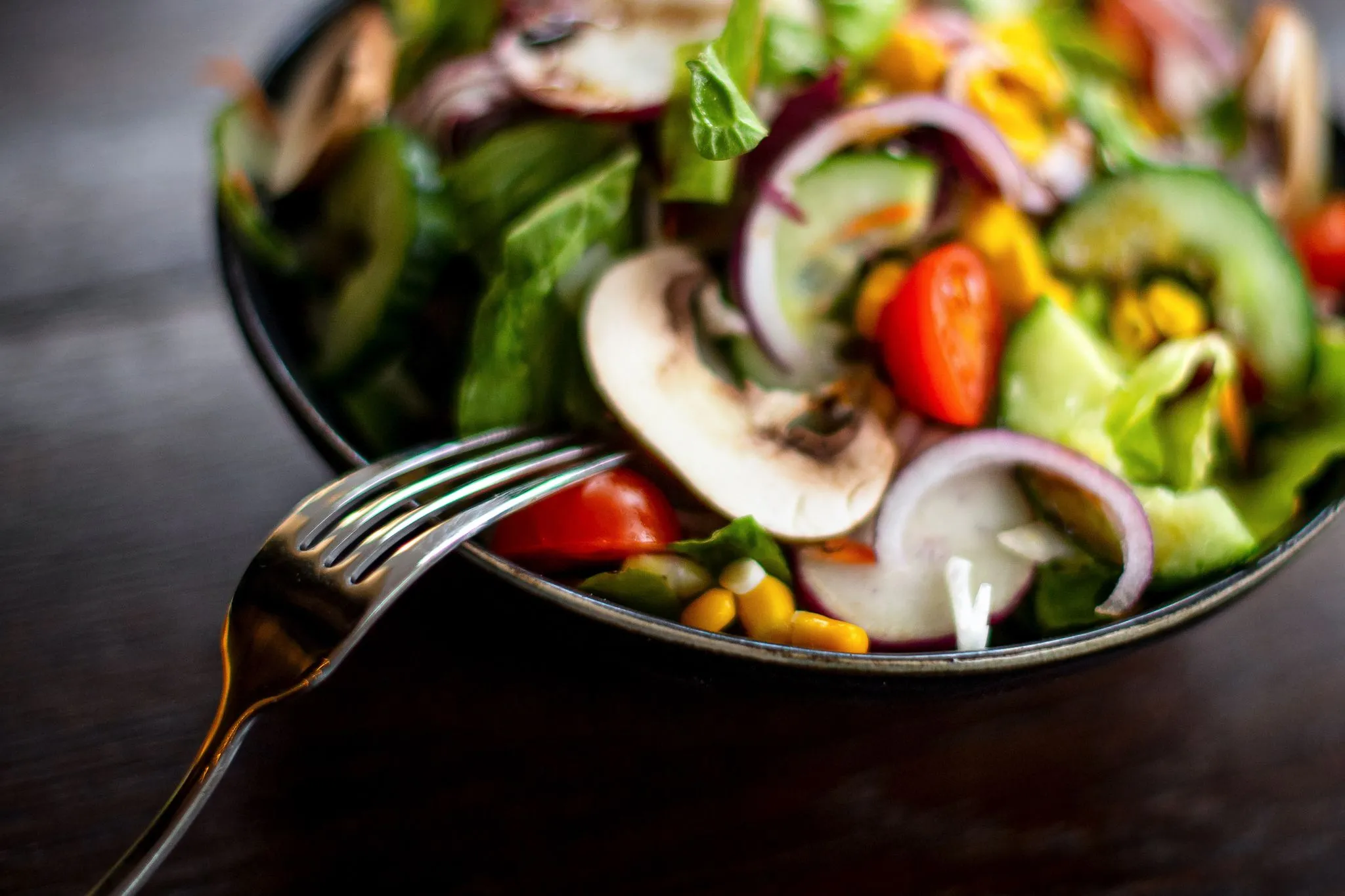 Close-up of a fresh salad with tomatoes, cucumbers, onions, and mushrooms in a dark bowl with a fork, vibrant colors, ap