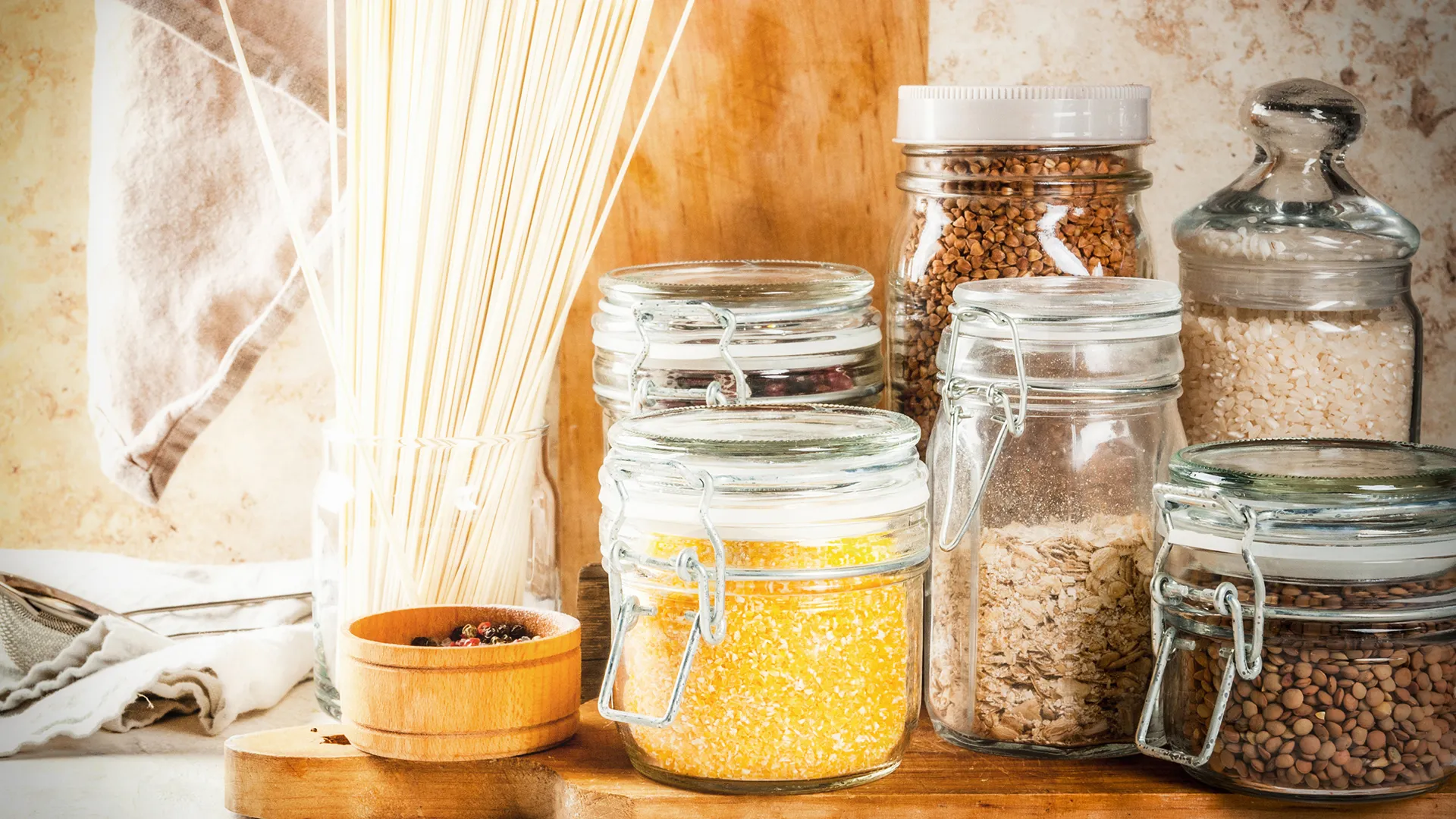 Glass jars filled with various grains, oats, lentils and dried noodles on a wooden counter, warm rustic mood