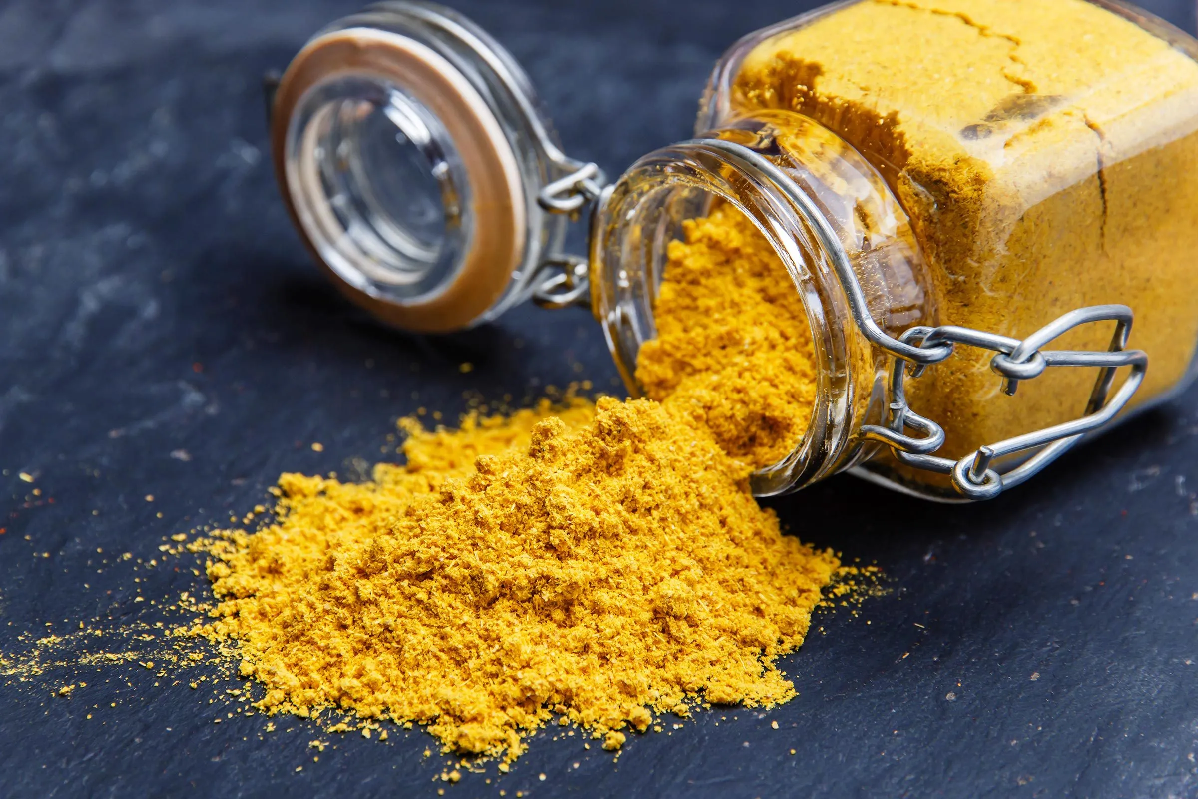Close-up of a glass jar tipped over spilling bright orange-yellow spice powder onto a dark surface, vivid warm colors