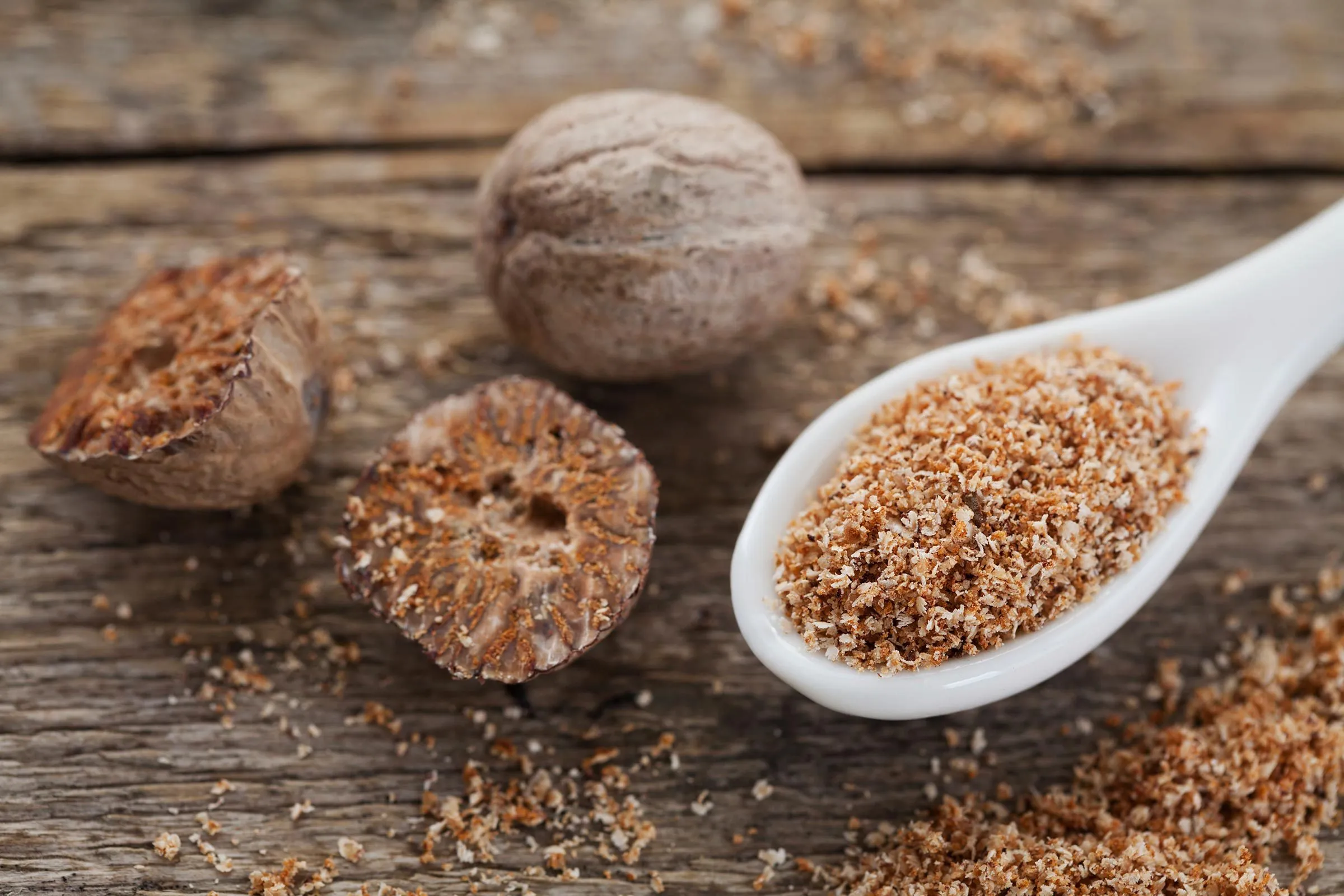 Close-up of whole and halved nutmeg seeds beside a white spoon filled with ground spice on a rustic wooden surface, warm
